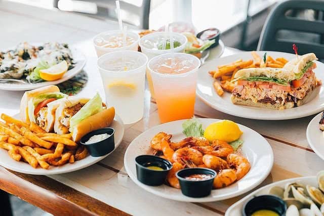 Plates on a table with shrimp, fries, sandwiches, drinks, and lemon wedges.