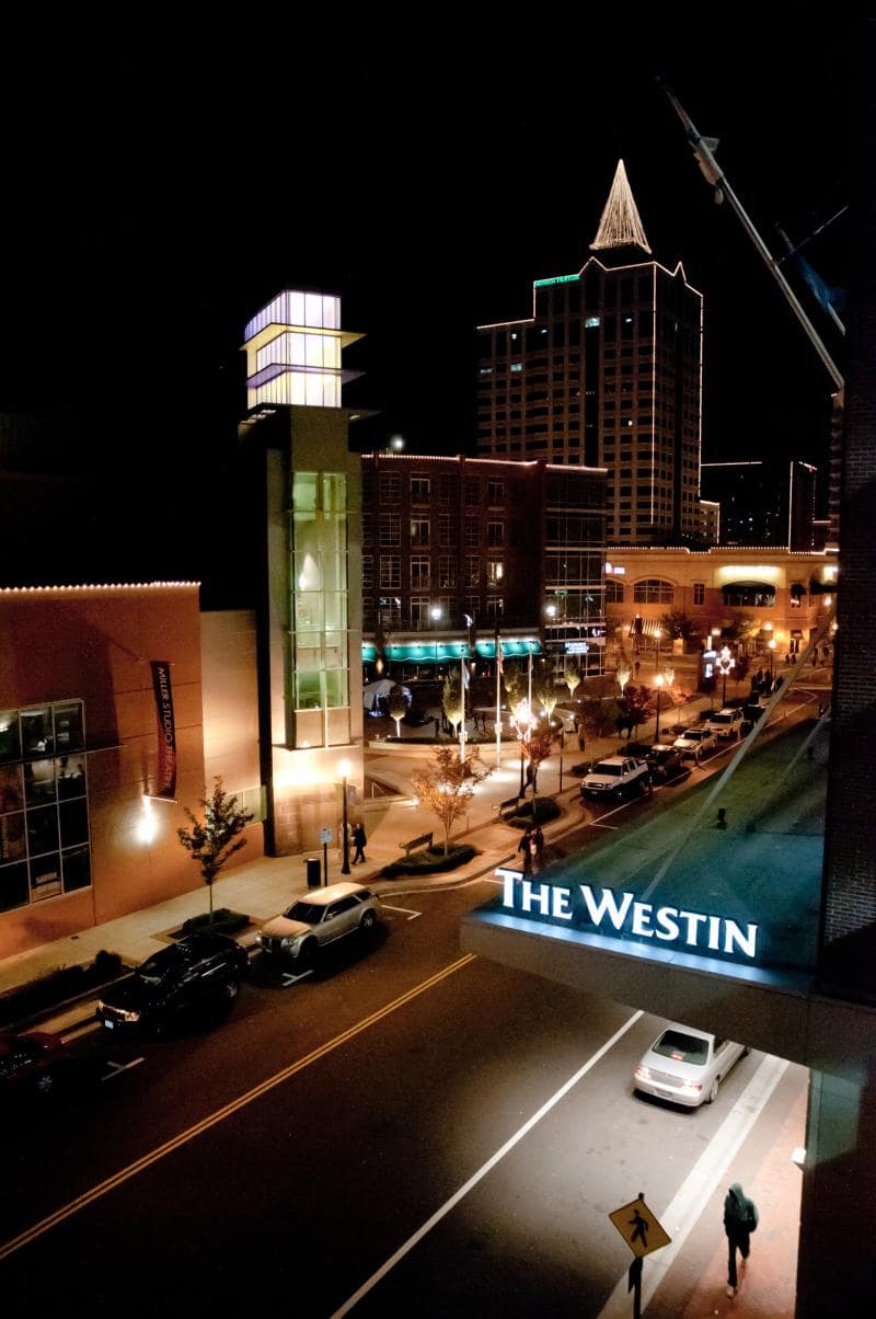 Night view of a city street with The Westin hotel entrance in the foreground, tall buildings in the background, and illuminated streetlights lining the road.