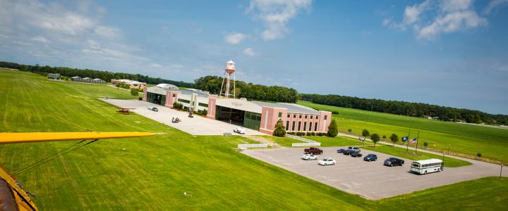 Aerial view of the Military Aviation Museum with a water tower, surrounded by green fields and a parking lot with cars and a bus.