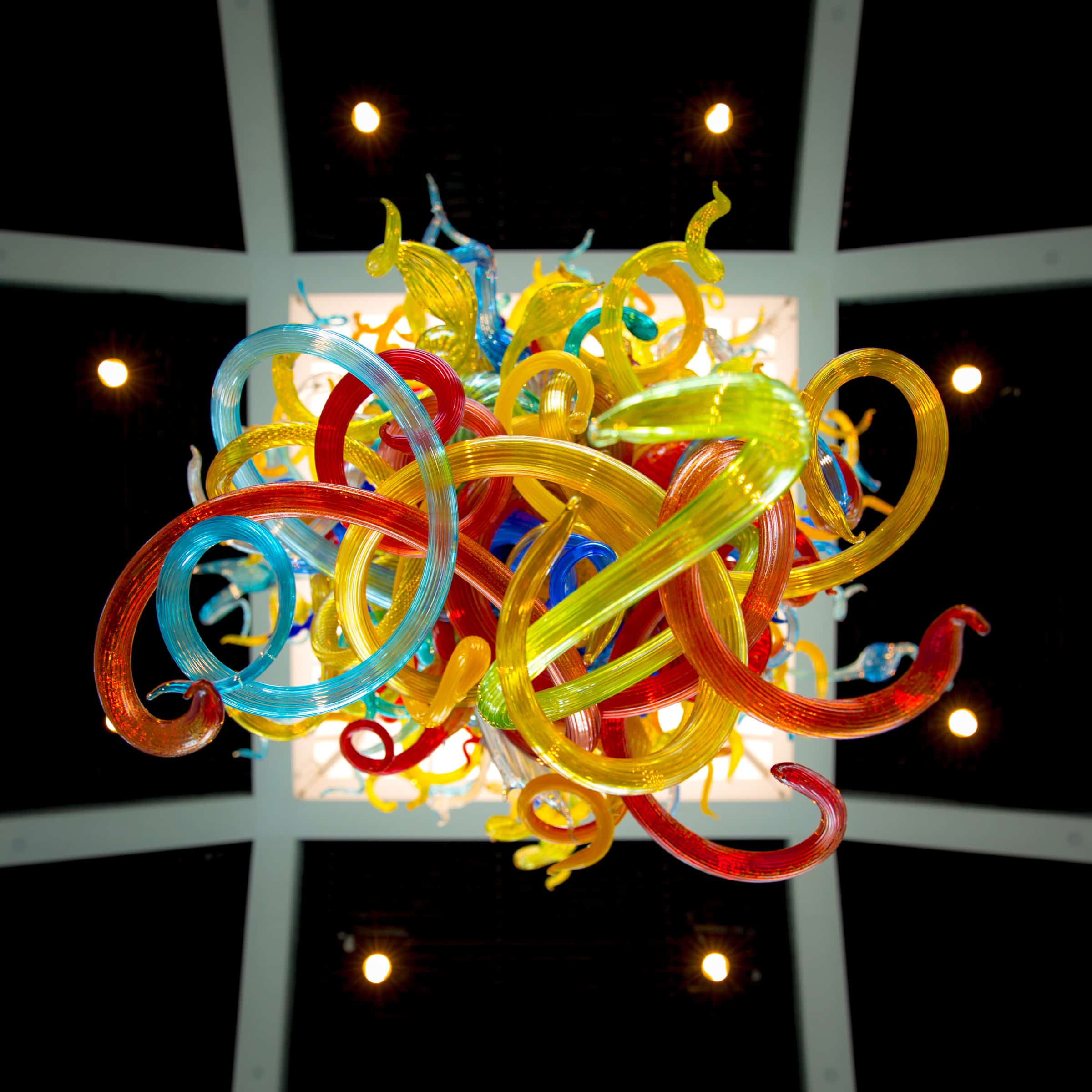 Colorful glass sculpture with intertwined red, yellow, and blue curves, viewed from below, set against a dark ceiling with lights.