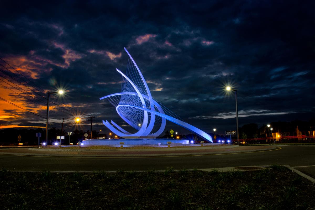 An illuminated, abstract steel sculpture stands in the center of a roundabout at dusk, with dramatic clouds and streetlights in the background.