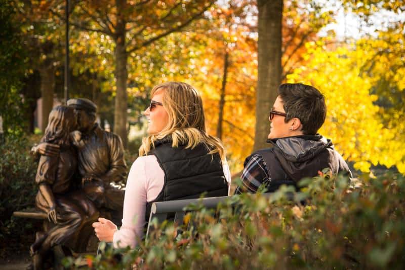 Two people sit on a park bench wearing sunglasses and vests, looking at a bronze statue of a couple seated nearby, with autumn trees in the background.