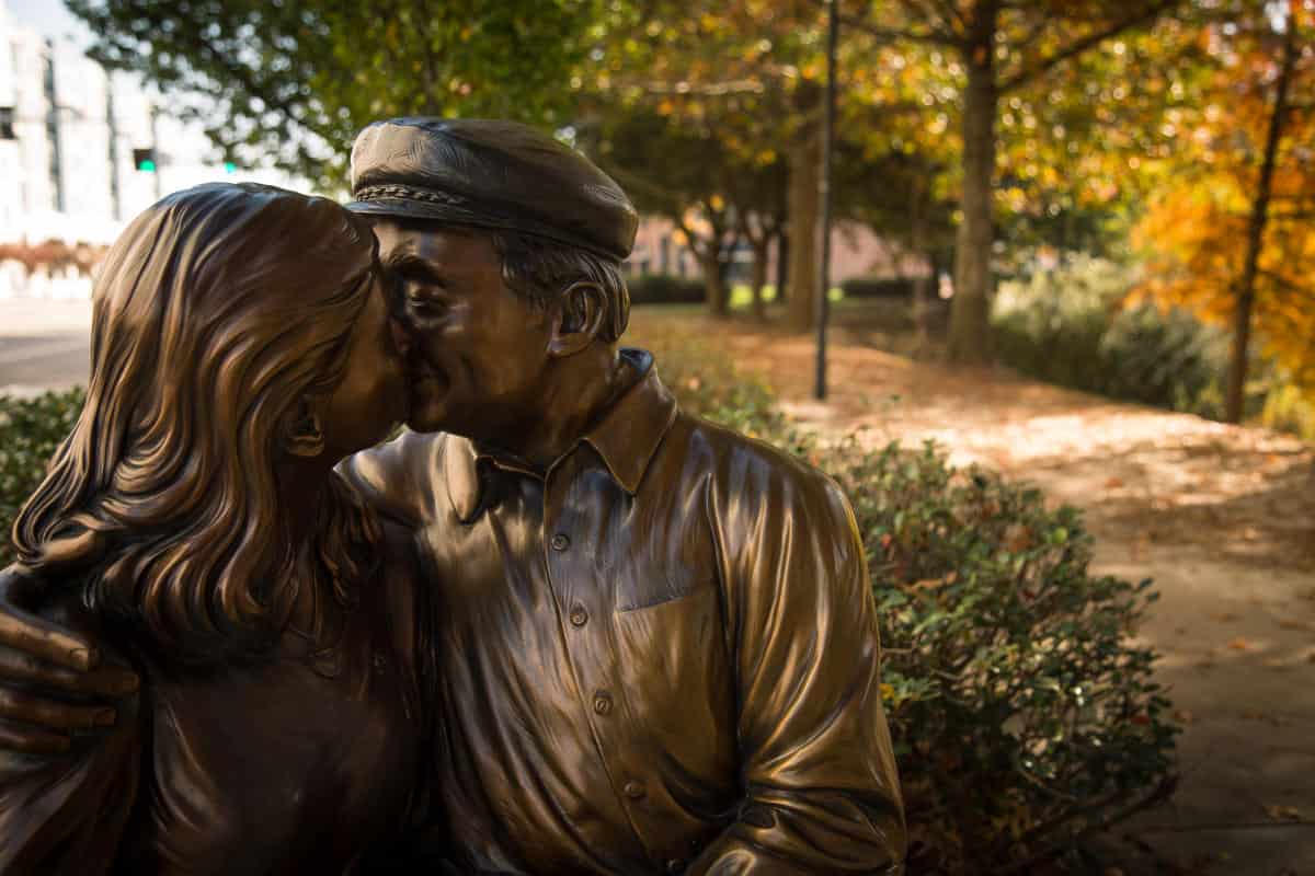 A bronze statue of a man in a cap and a woman with long hair sharing a kiss, set in an outdoor park with trees and a pathway in the background.