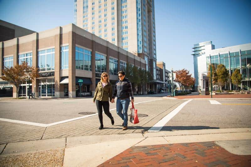 A couple walks hand in hand across an intersection in a city area, with a tall building and shops in the background.