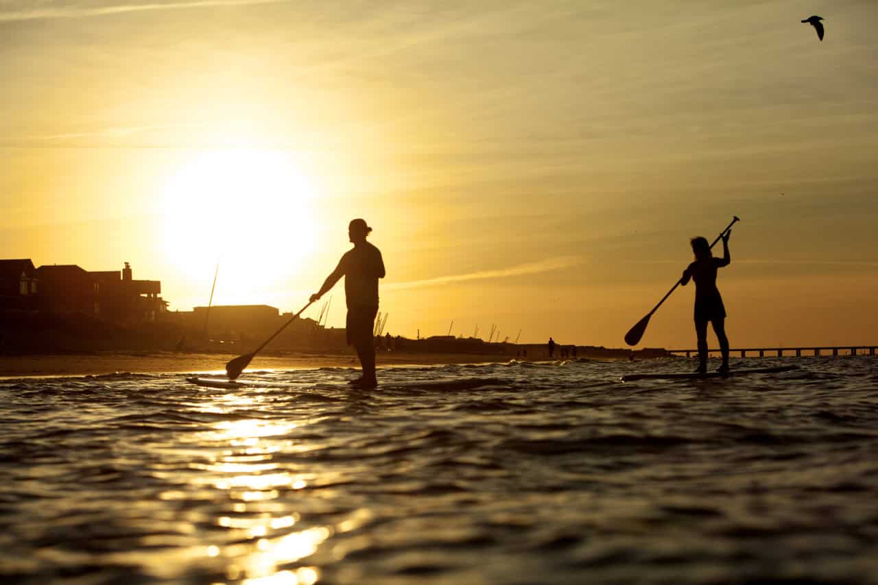 Two people are paddleboarding on the water during sunset, with a bird flying overhead and structures visible on the shore.