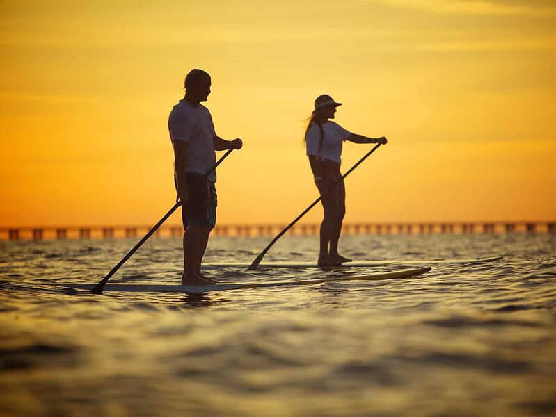 Two people paddleboarding on calm water during sunset, with a silhouette of a bridge in the background.