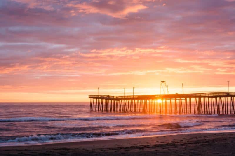 Sunset view over an ocean pier with waves gently crashing on the shore, under a vibrant sky of pink and orange hues.