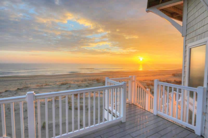 Sunset view from a beach house deck with white railing, overlooking a sandy beach and ocean under a partly cloudy sky.