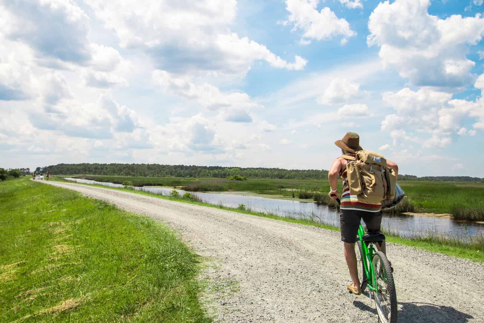 Person cycling on a gravel road beside a grassy field and waterway, under a partly cloudy sky, with a backpack and hat.