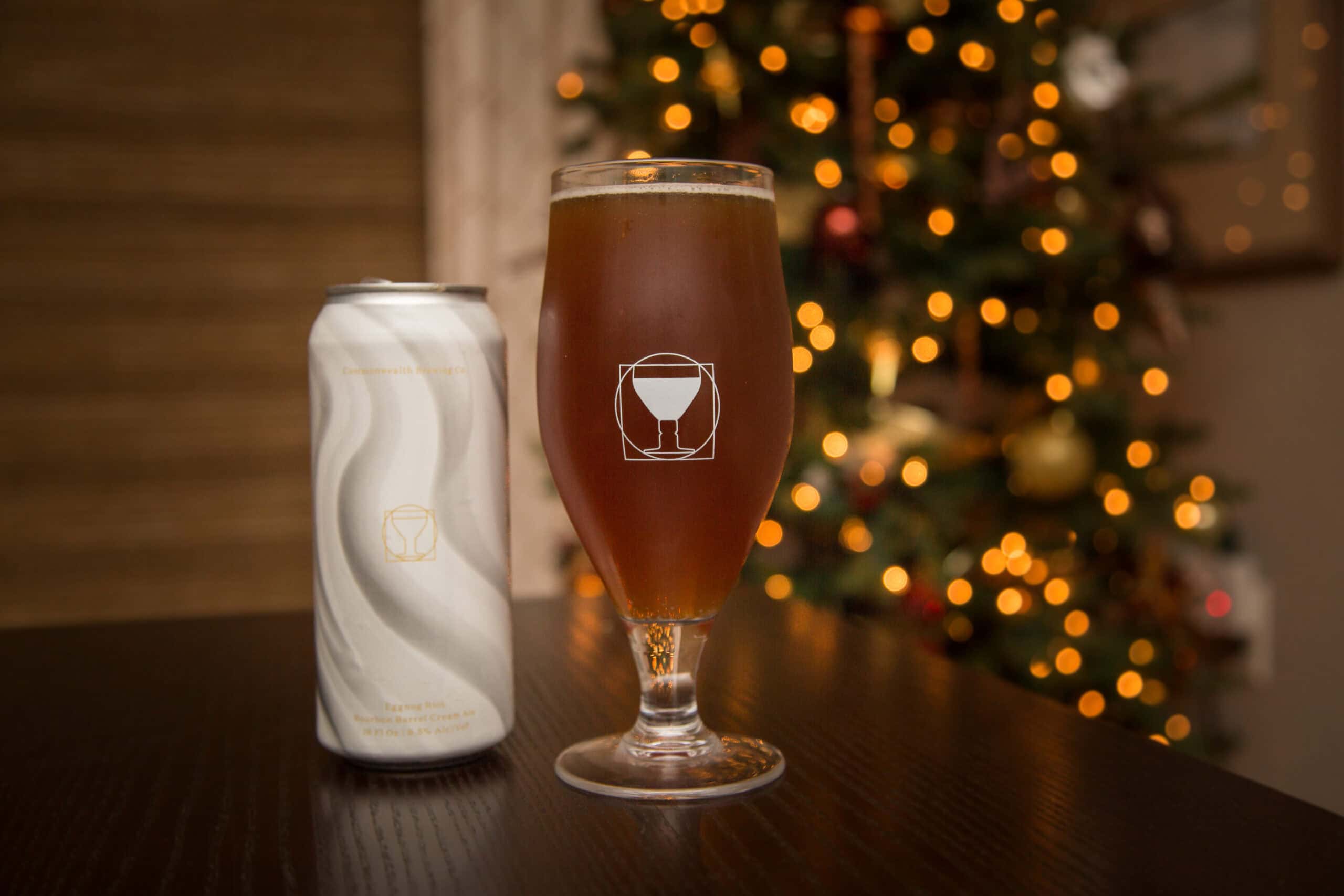 A beer can and a glass of beer sit on a dark table in front of a blurred, lit Christmas tree.