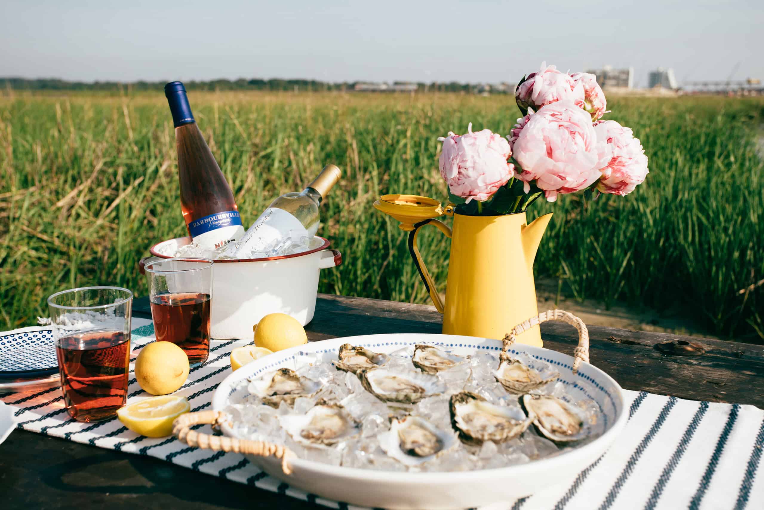 A picnic setup with oysters on ice, lemon halves, bottles of wine, glasses with red drink, and pink flowers in a yellow pitcher, set on a striped cloth in a grassy field.