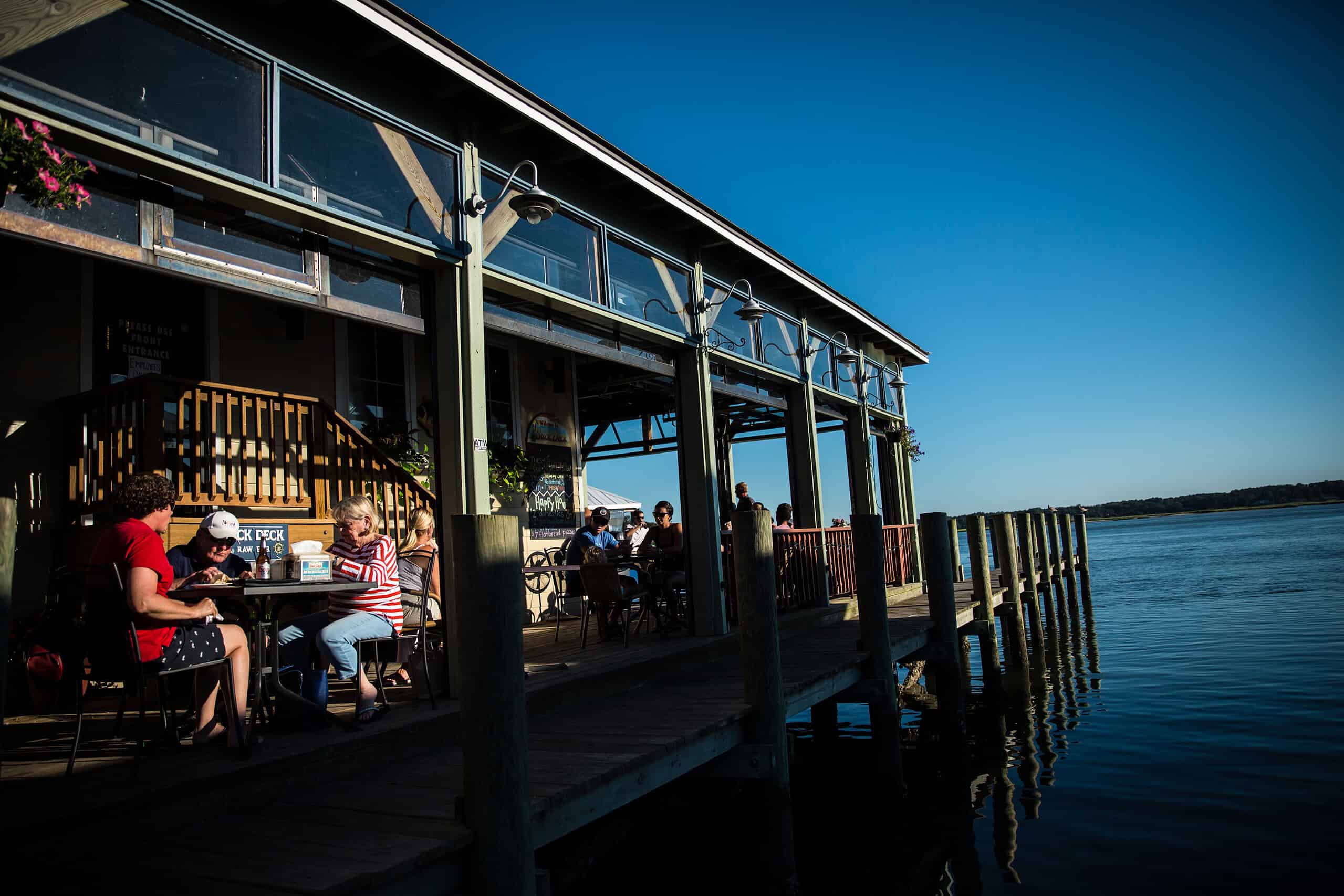 People are dining outdoors on a dockside restaurant under a clear blue sky, with a view of the water on the right.