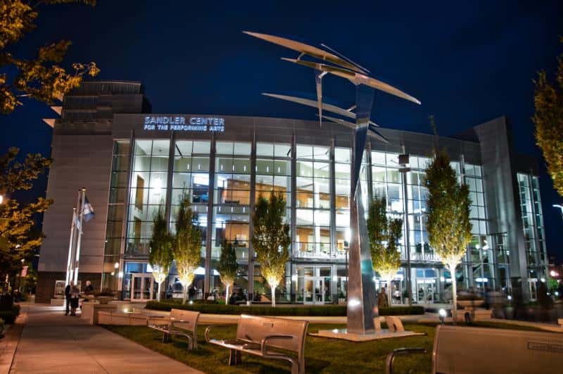 Exterior of the Sandler Center for the Performing Arts at night, with illuminated glass facade and a modern sculpture in the foreground. Benches and trees line the walkway.
