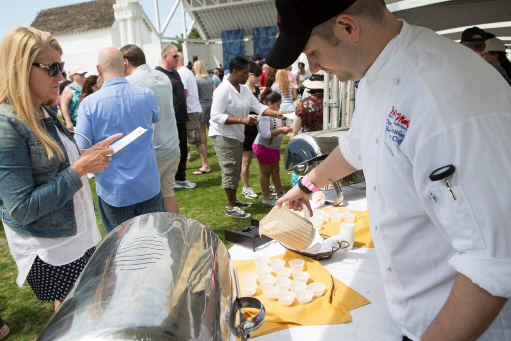 A chef in a white jacket pours a beverage from a pitcher into small cups at a busy outdoor event, while attendants look on.