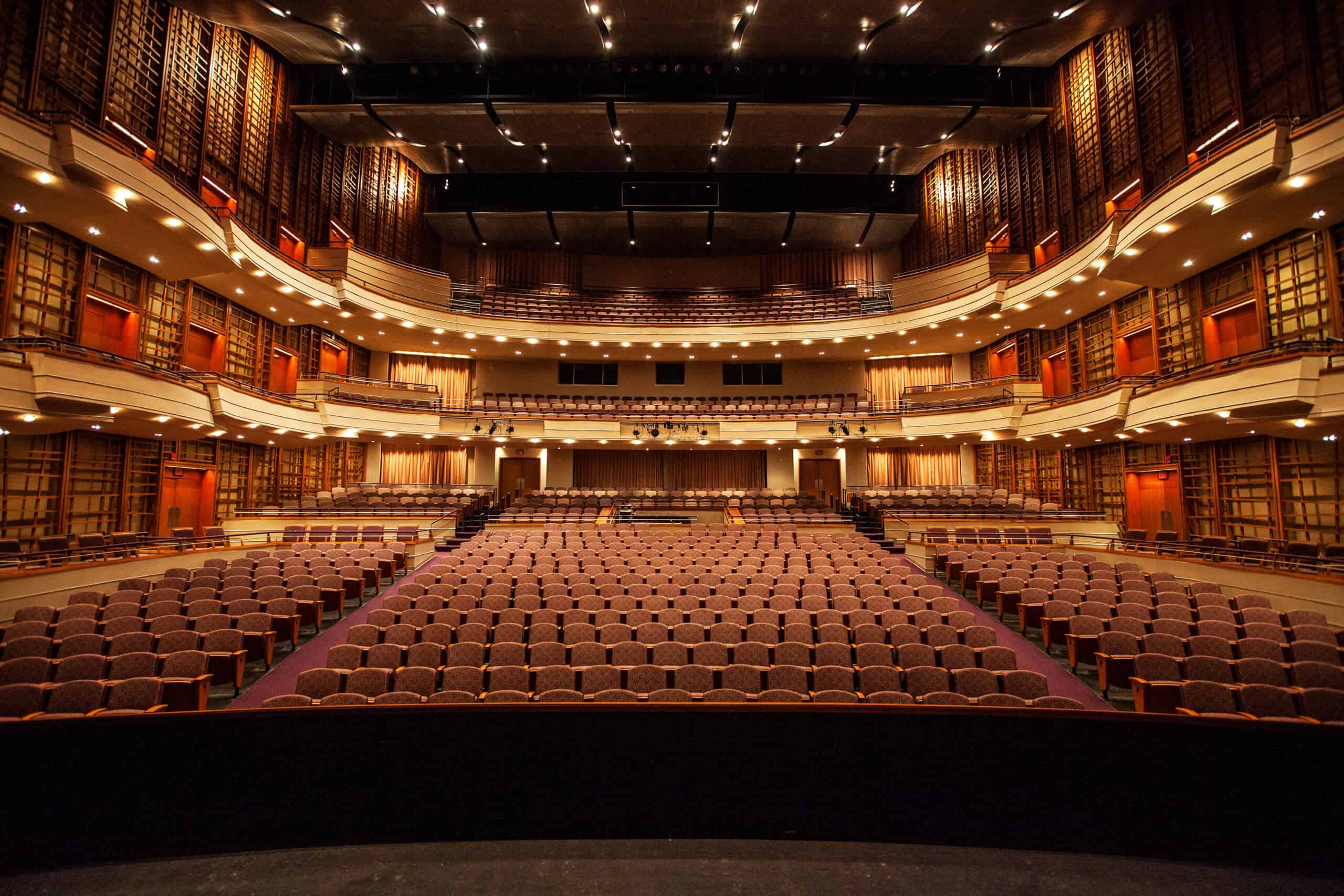 Interior of an empty theater auditorium with rows of brown seats, multiple balconies, and warm lighting. Stage view facing the audience.