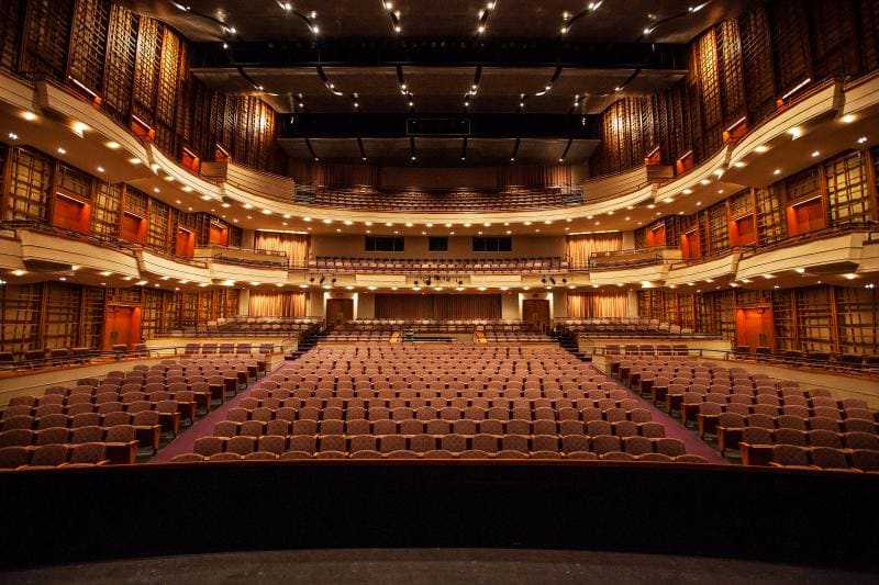 Empty, well-lit concert hall interior with multiple seating tiers and a clear view of the stage from the audience's perspective.