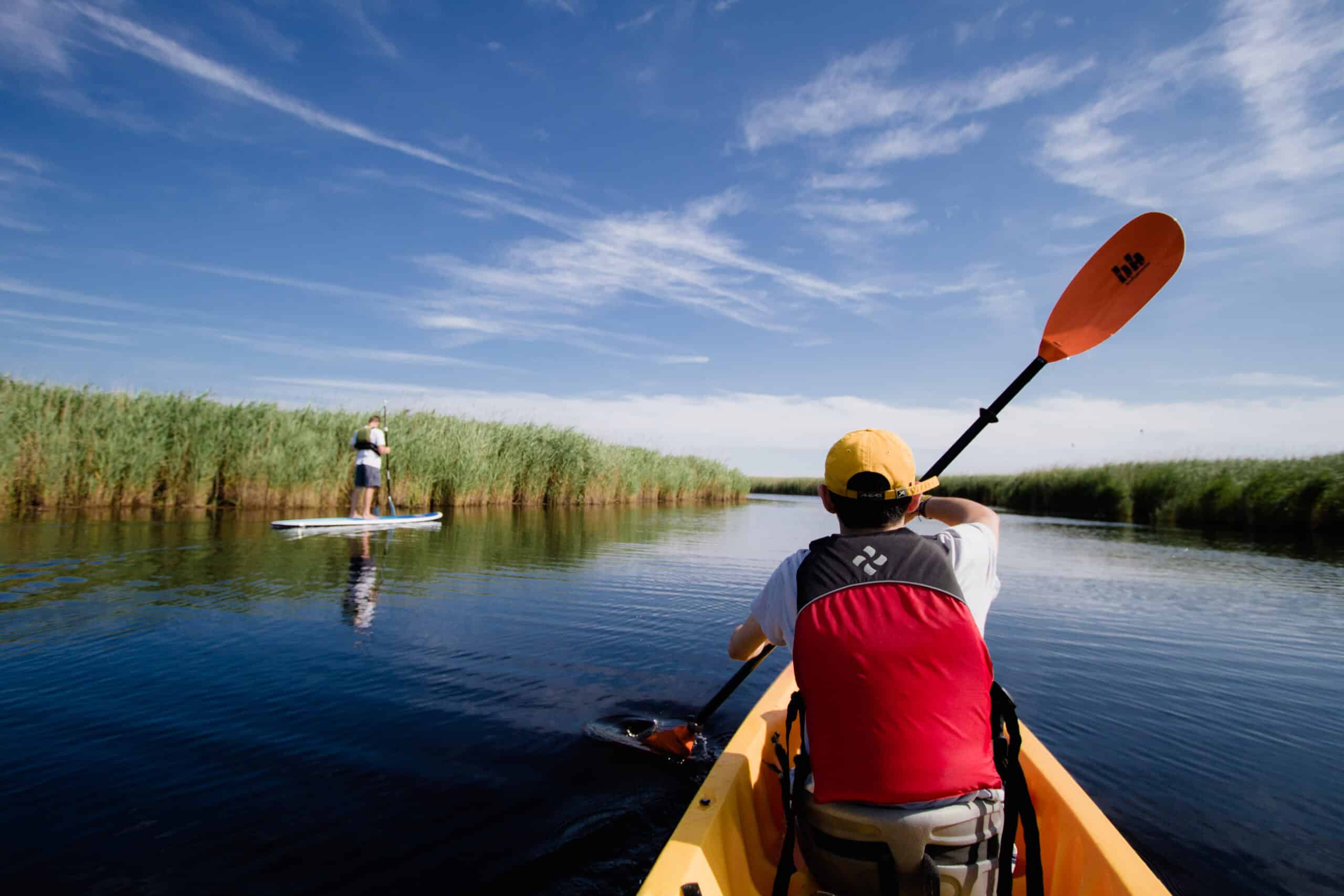 Person kayaking in a waterway surrounded by tall grass, with another person paddleboarding ahead under a clear blue sky.