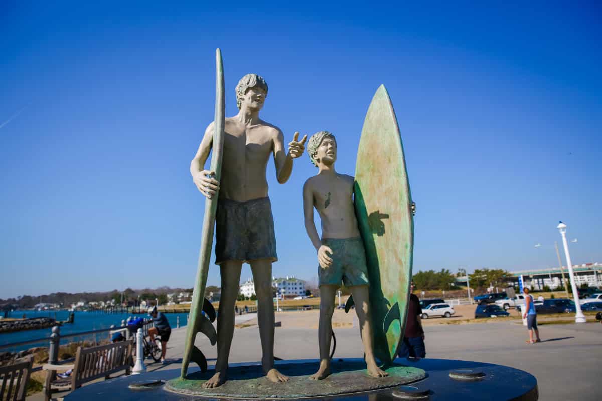 Statue of two surfers with surfboards standing on a circular base at the Virginia each oceanfront under a clear blue sky.