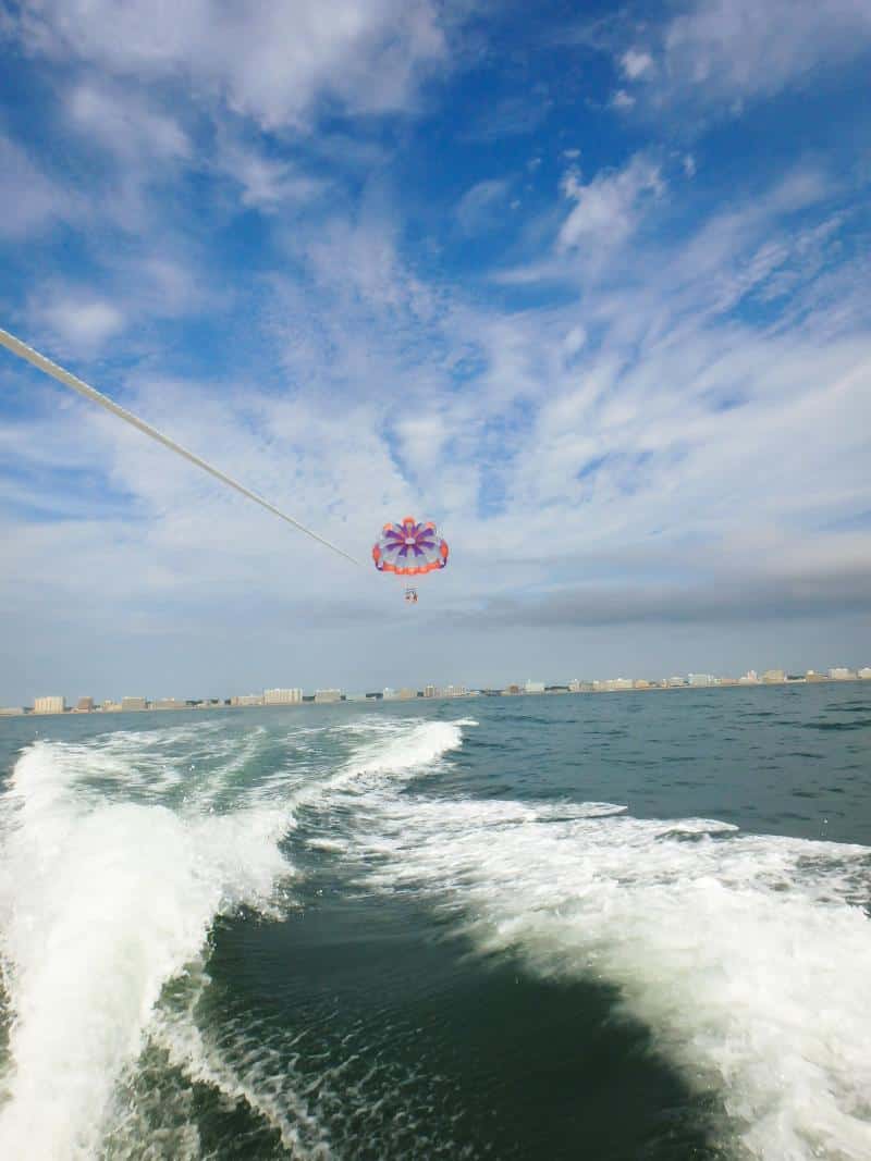 A person parasailing over the ocean, attached to a parachute, with a view of waves and a distant city skyline under a partly cloudy sky.