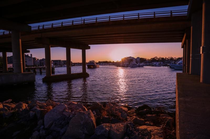 Sunset view from under a bridge, overlooking a waterway with boats in the distance.