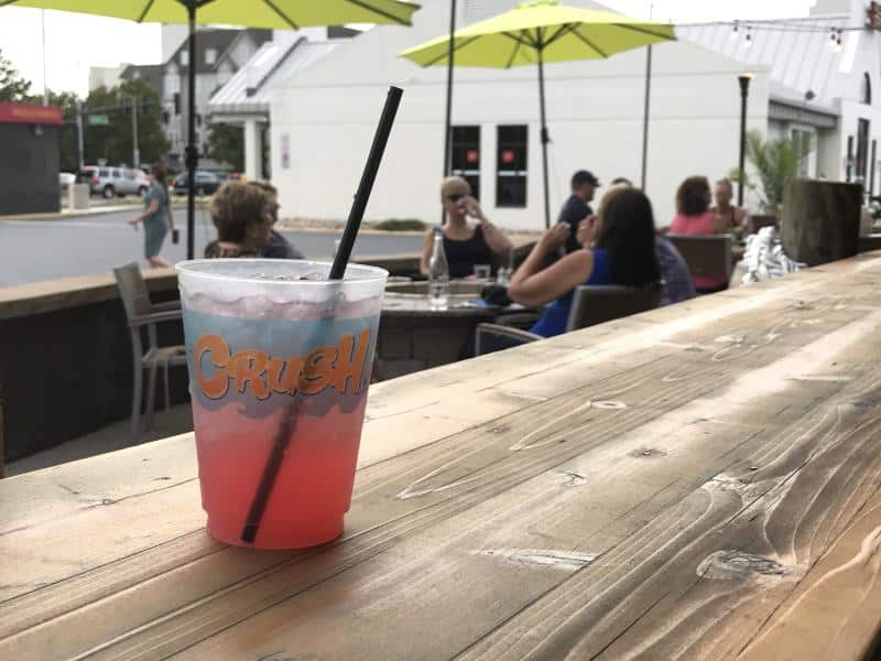 A pink drink in a plastic cup with "Crush" printed on it sits on a wooden table outdoors, with people sitting at tables with umbrellas in the background.
