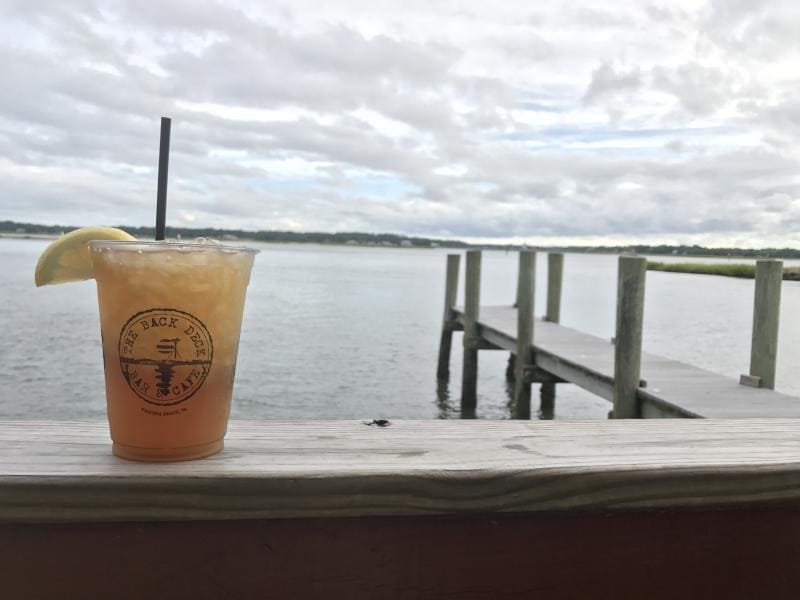 A plastic cup of iced tea with a lemon slice and straw sits on a wooden railing overlooking a cloudy lake and a dock.