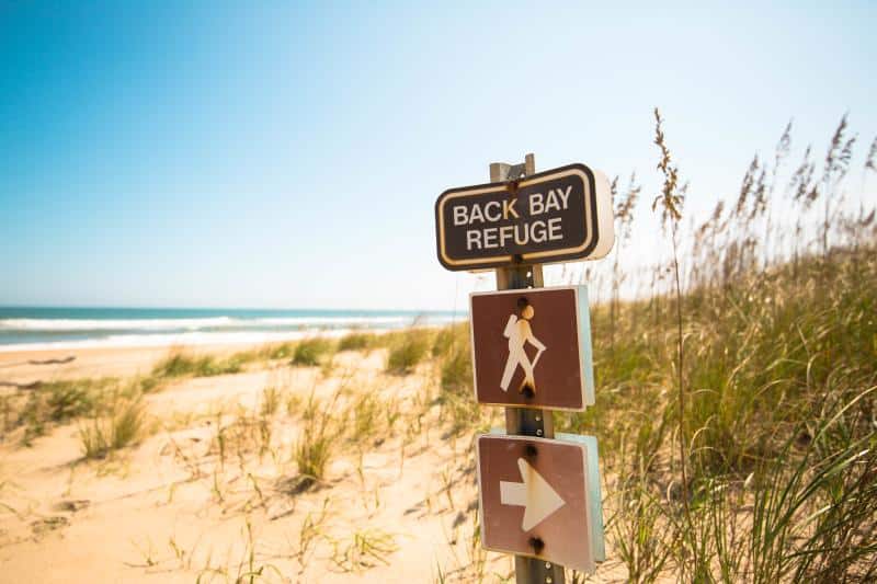 Signpost for Back Bay Refuge with walking and arrow symbols, set on a sandy beach with grass and ocean in the background.
