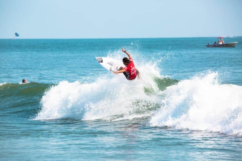 A surfer in a red shirt performs a jump on a wave in the ocean, with a boat visible in the background.