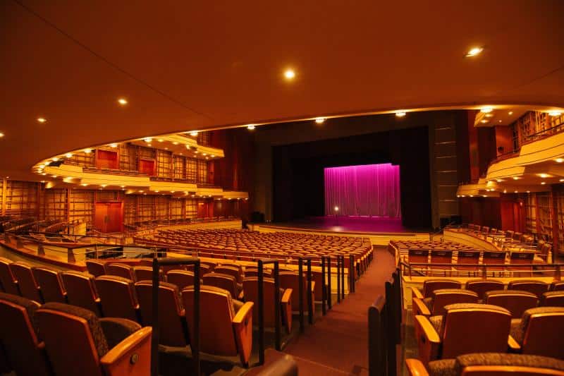 Empty theater with rows of brown seats facing a stage with a closed pink curtain. Curved balcony and walls with wood paneling create an elegant atmosphere.