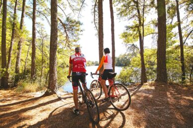 A man and woman biking on a path in the woods overlooking freshwater with sunlight glimmering through the trees.