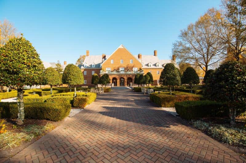 A large brick mansion with arched entrance and multiple chimneys is surrounded by a formal garden with neatly trimmed hedges and trees under a clear blue sky.