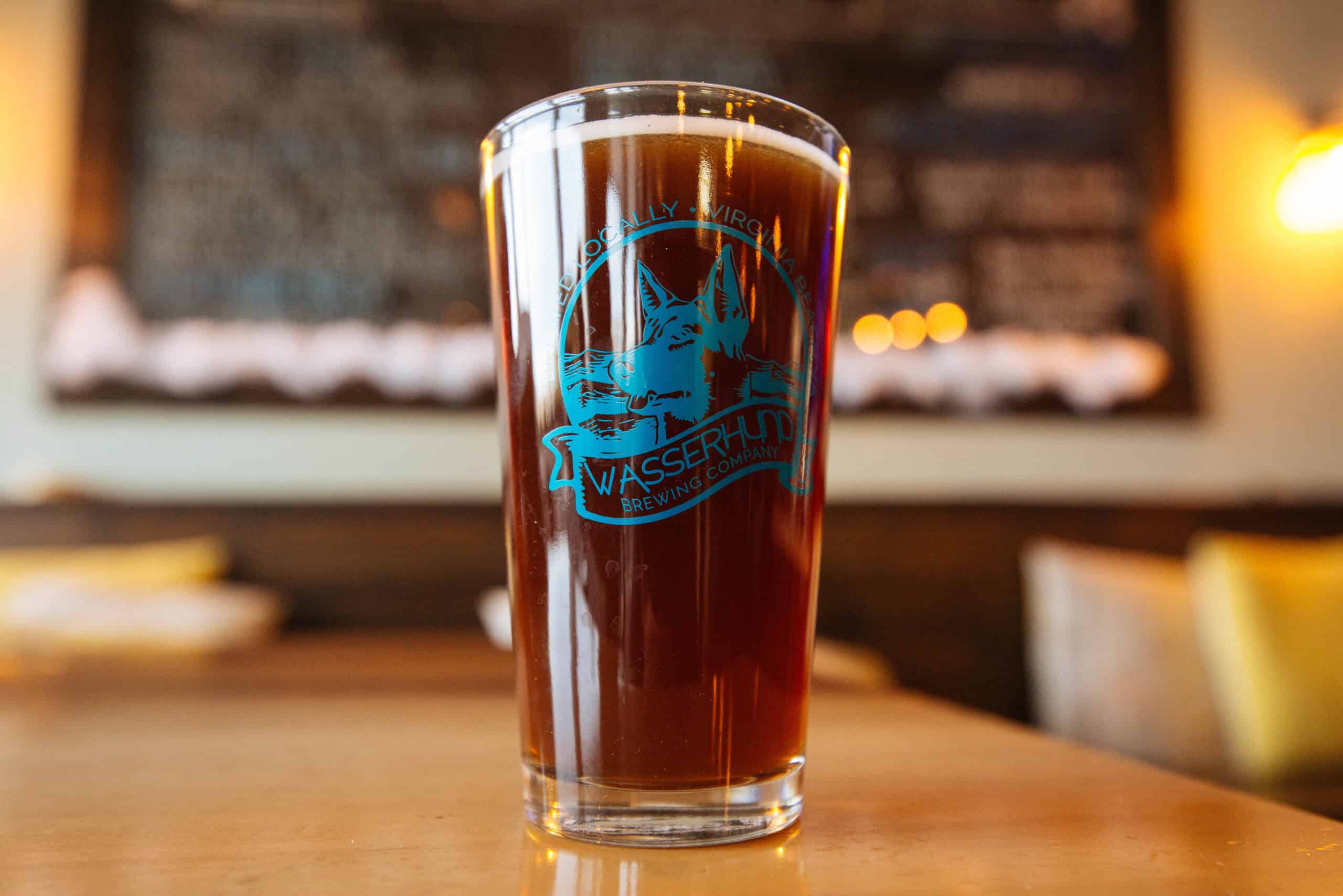 A pint glass filled with dark beer sits on a wooden table. The glass has a blue logo for "Wasserhund Brewing Company." Blurred background with soft lighting.