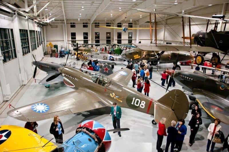 Aircraft museum with various vintage airplanes on display. People walk around the exhibits inside a large hangar.