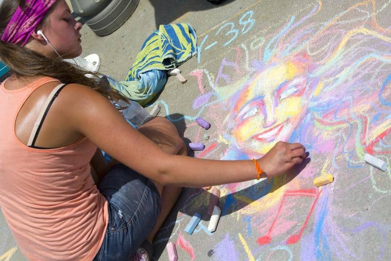 Person drawing a colorful chalk art of a smiling face on the sidewalk, with multicolored chalks scattered around.