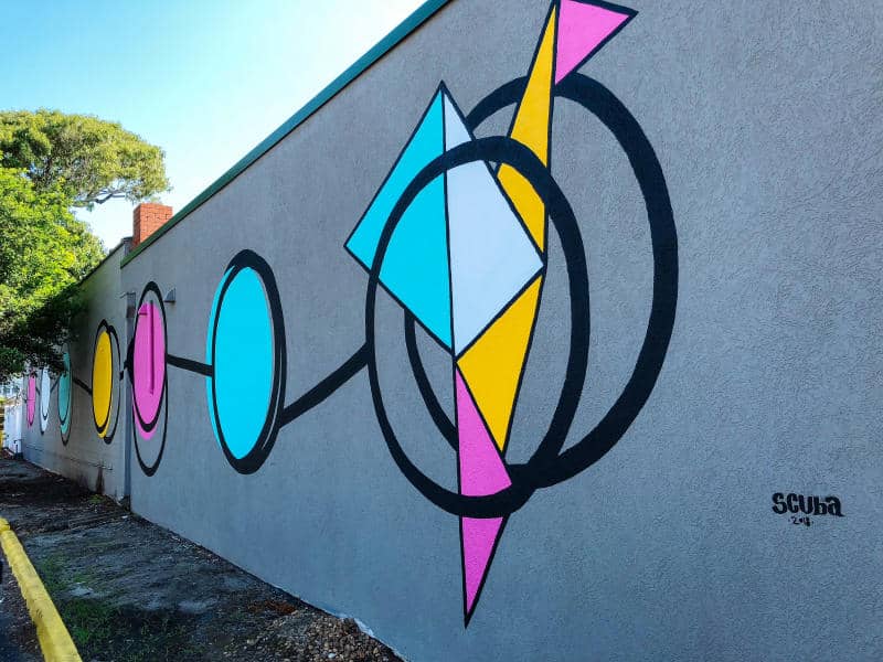Geometric mural with circles, triangles, and rectangles in blue, pink, yellow, and black on a gray wall.