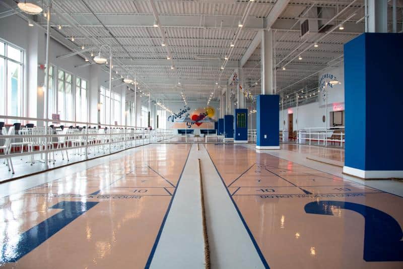 Indoor shuffleboard court with polished floor and numbered markings, surrounded by tables and chairs under a high ceiling with exposed beams.