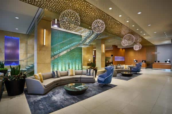 Modern hotel lobby with elegant decor, featuring a curved sofa, blue chairs, spherical chandeliers, a glass staircase, and a water feature on the wall. Reception desks in the background.