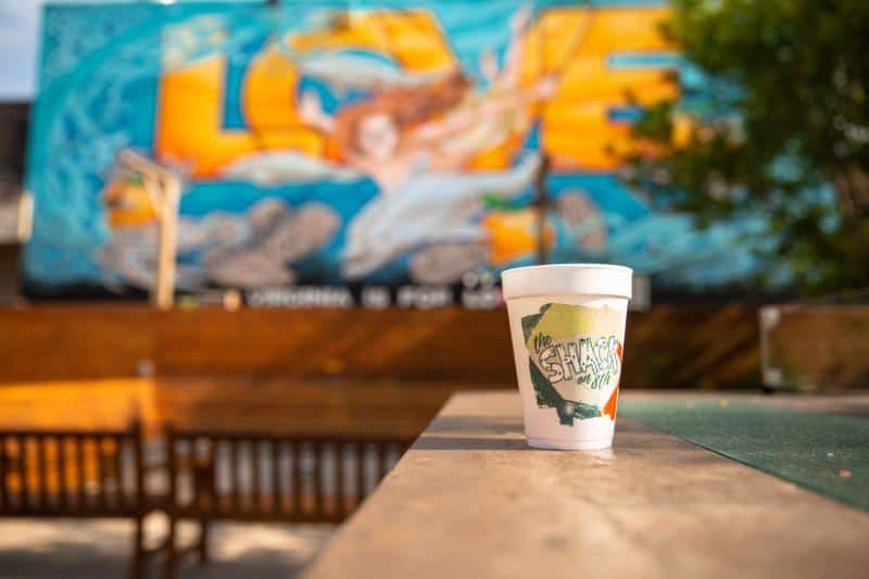 A white cup with colorful graffiti design sits on a table outdoors. A vibrant mural with the word "LOVE" and a woman is in the background, with a bench and trees nearby.