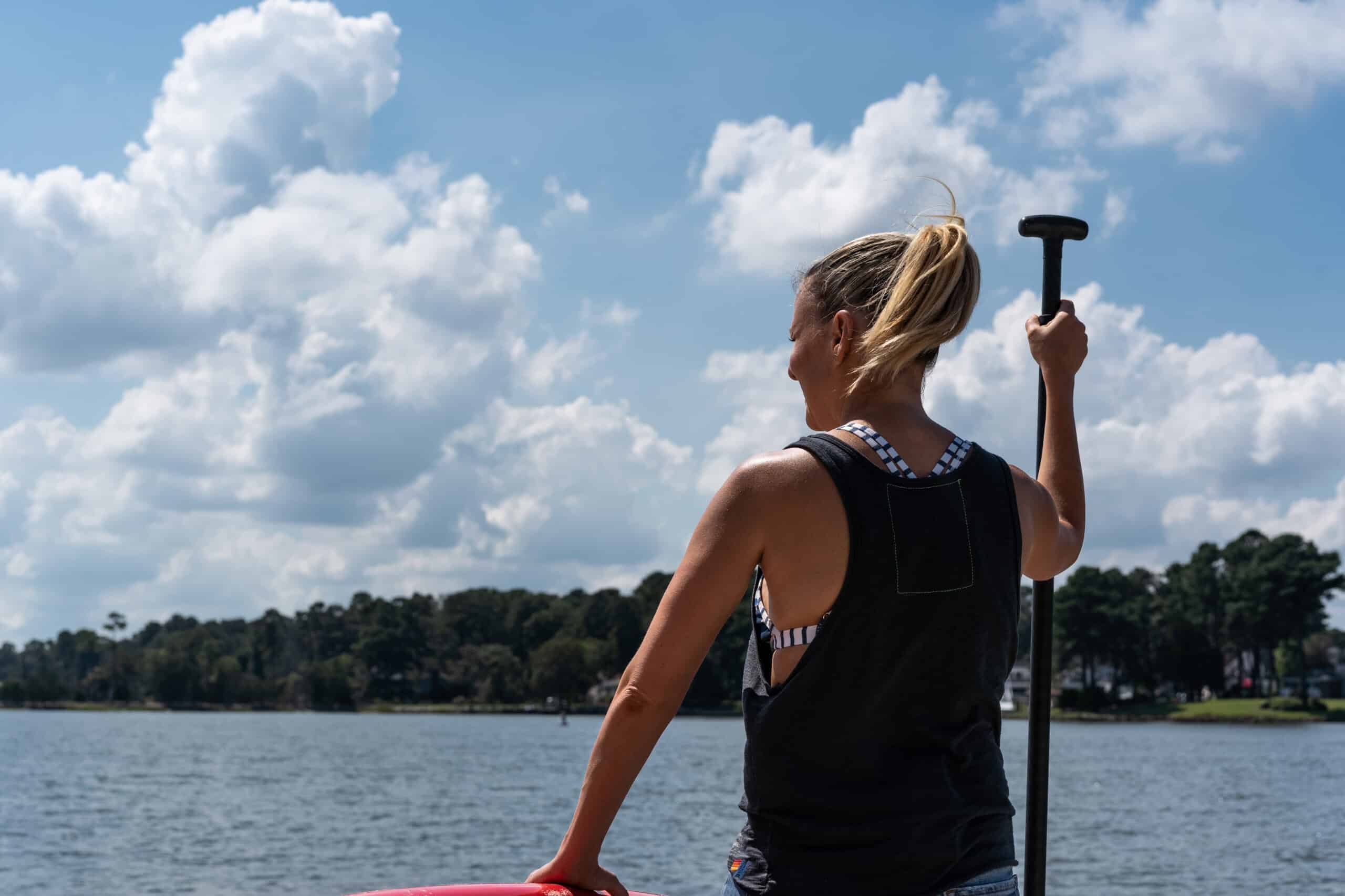 A woman with a ponytail stands on a paddleboard holding a paddle, looking out at a lake with trees in the background under a partly cloudy sky.