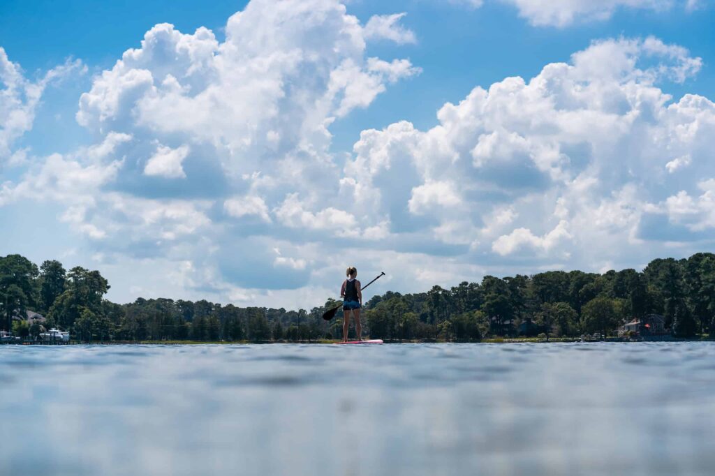 A person is paddleboarding on a calm lake under a partly cloudy sky, with trees lining the distant shore.