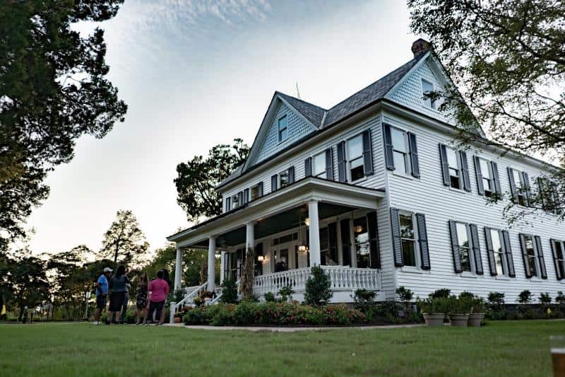 A group of people stands in front of a large, white, two-story house with a porch and gabled roof, surrounded by trees and green lawn, under a clear sky.