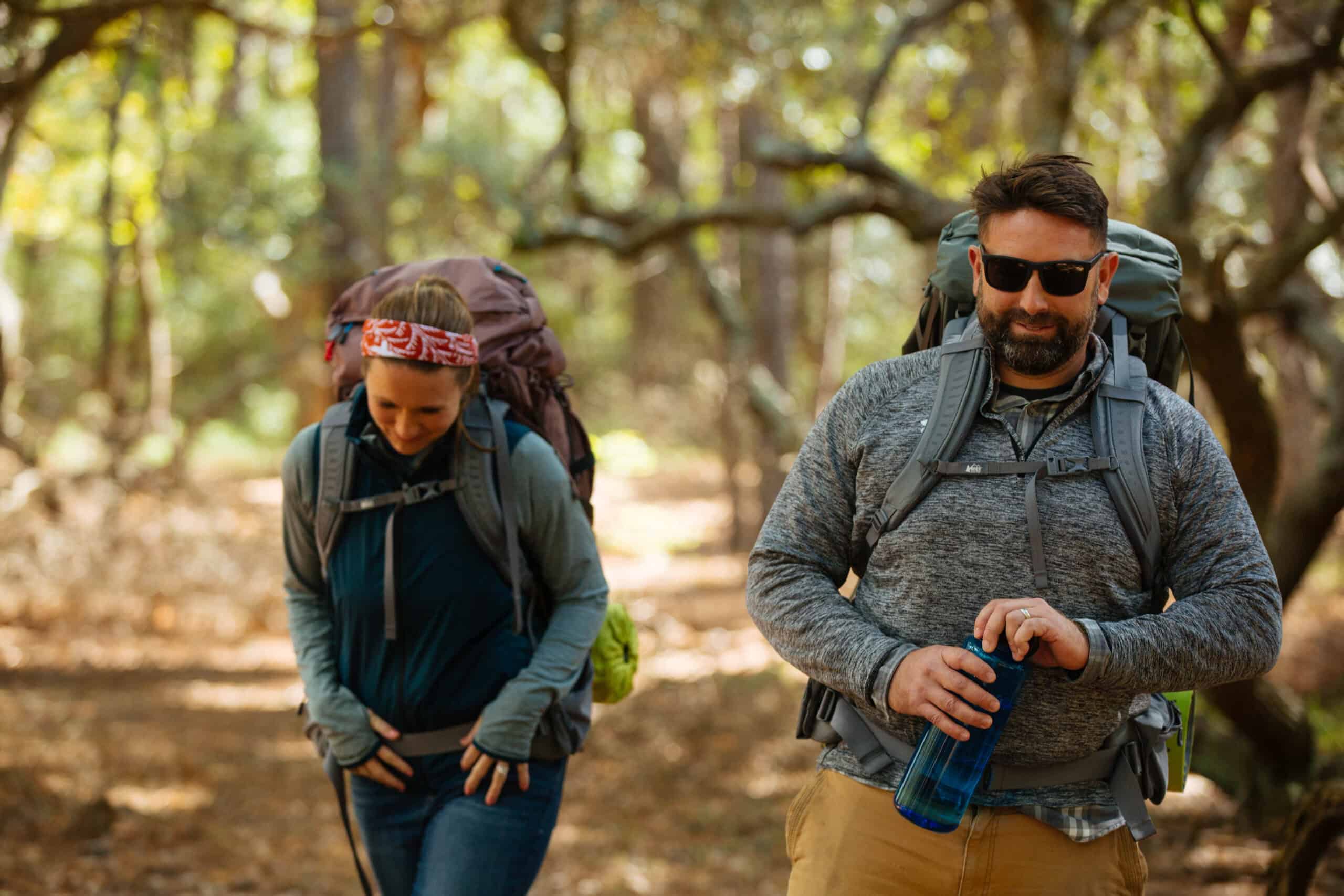 Two people with backpacks walk through a forested trail. The man holds a blue water bottle, and the woman wears a headband. Sunlight filters through the trees.