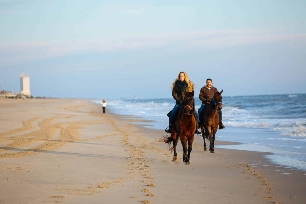 Two people ride horses along a sandy beach with ocean waves and a distant figure walking.