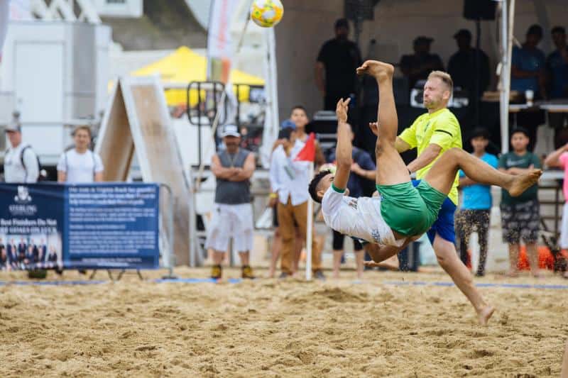 Two men playing beach soccer, one in mid-air attempting a bicycle kick. Spectators and a referee watch from the sidelines.