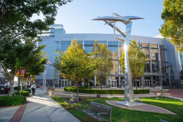 Modern building exterior with large windows and a metal sculpture nearby. Trees and benches line the sidewalk. Sunlight filters through the leaves.
