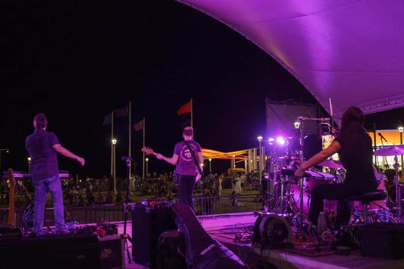 A band performs on an outdoor stage at night, with purple lighting. Musicians include a singer, guitarist, and drummer. An audience is visible in the background.