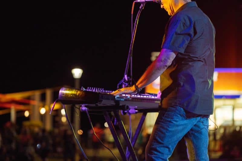 A musician plays a keyboard on a stand under stage lights at night, with a crowd blurred in the background.