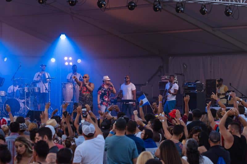 A band performs on stage under blue lighting at an outdoor concert with a large, cheering crowd in front.