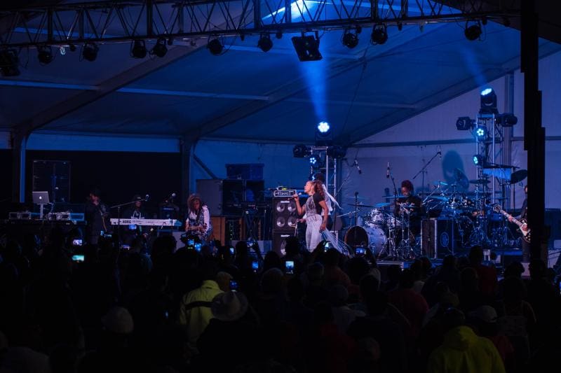 A band performs on an outdoor stage under a tent with blue lighting, surrounded by an audience.