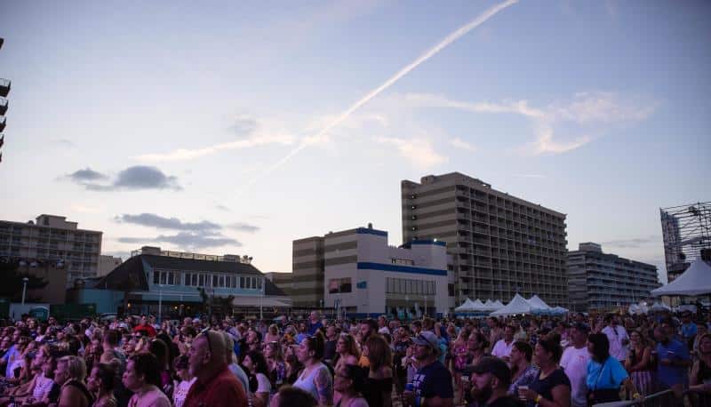 A crowd gathers at an outdoor event during sunset. Tall buildings and tents are visible in the background under a partly cloudy sky.